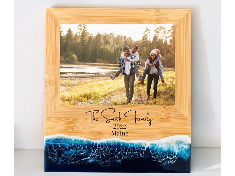 May include: A wooden photo frame with a blue and white ocean wave design at the bottom. The frame has a photo of a family of four in a field. The text "The Smith Family 2022 Maine" is inscribed on the frame.