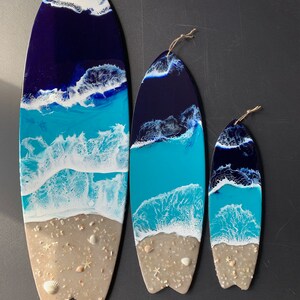 May include: Three surfboard-shaped resin art pieces with a beach scene. The largest piece is blue, white, and tan with a blue ocean, white waves, and tan sand. The medium piece is the same design but smaller. The smallest piece is the same design but even smaller.