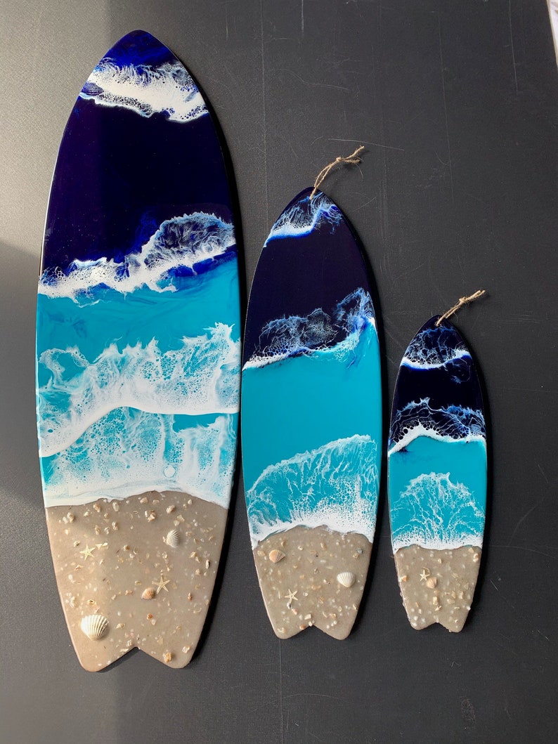 May include: Three surfboard-shaped resin art pieces with a beach scene. The largest piece is blue, white, and tan with a blue ocean, white waves, and tan sand. The medium piece is the same design but smaller. The smallest piece is the same design but even smaller.
