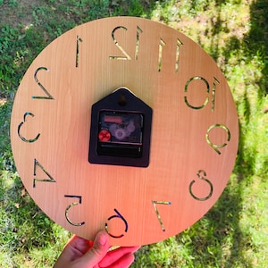 May include: A wooden clock face with Roman numeral hour markers. The clock face is light brown and the numbers are black. The clock is held in a hand.