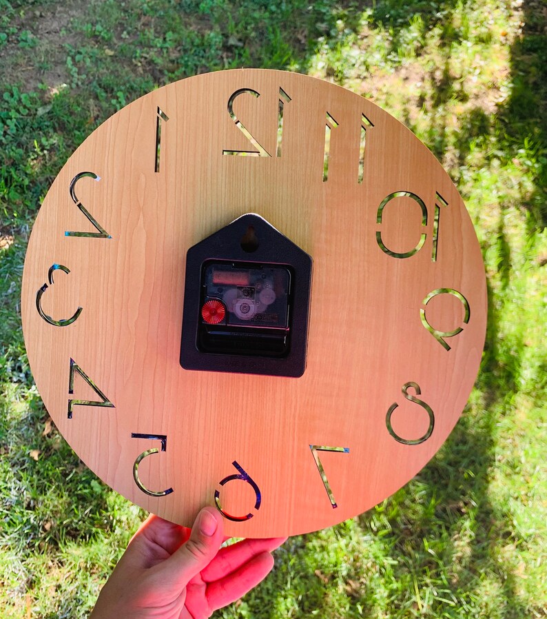 May include: A wooden clock face with Roman numeral hour markers. The clock face is light brown and the numbers are black. The clock is held in a hand.