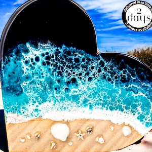 May include: A heart-shaped resin art piece depicting an ocean scene with blue and white waves crashing on a sandy beach. The piece is set against a black background and includes small white seashells and a starfish.