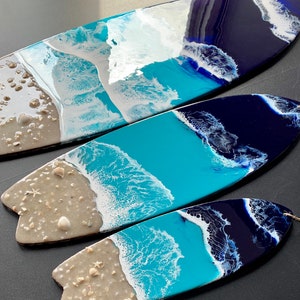 May include: Three surfboard-shaped resin art pieces with a beach theme. The pieces are painted with shades of blue, white, and gray to represent the ocean, waves, and sand. The largest piece has a more abstract design, while the smaller pieces have a more realistic depiction of the beach.