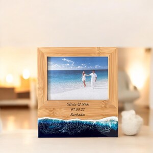 May include: A bamboo photo frame with a blue and white resin wave design at the bottom. The frame has a photo of a couple walking on a beach and the text "Olivia & Nick 07.09.22 Barbados" engraved on the wood.