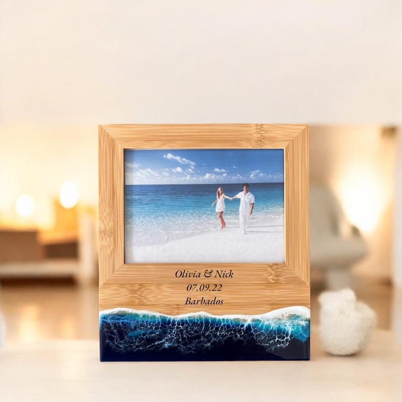 May include: A bamboo photo frame with a blue and white resin wave design at the bottom. The frame has a photo of a couple walking on a beach and the text "Olivia & Nick 07.09.22 Barbados" engraved on the wood.