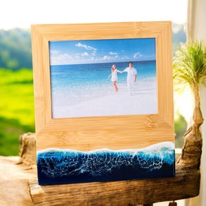 May include: A wooden picture frame with a blue and white resin base that resembles ocean waves. The frame holds a photo of a couple walking on a beach.