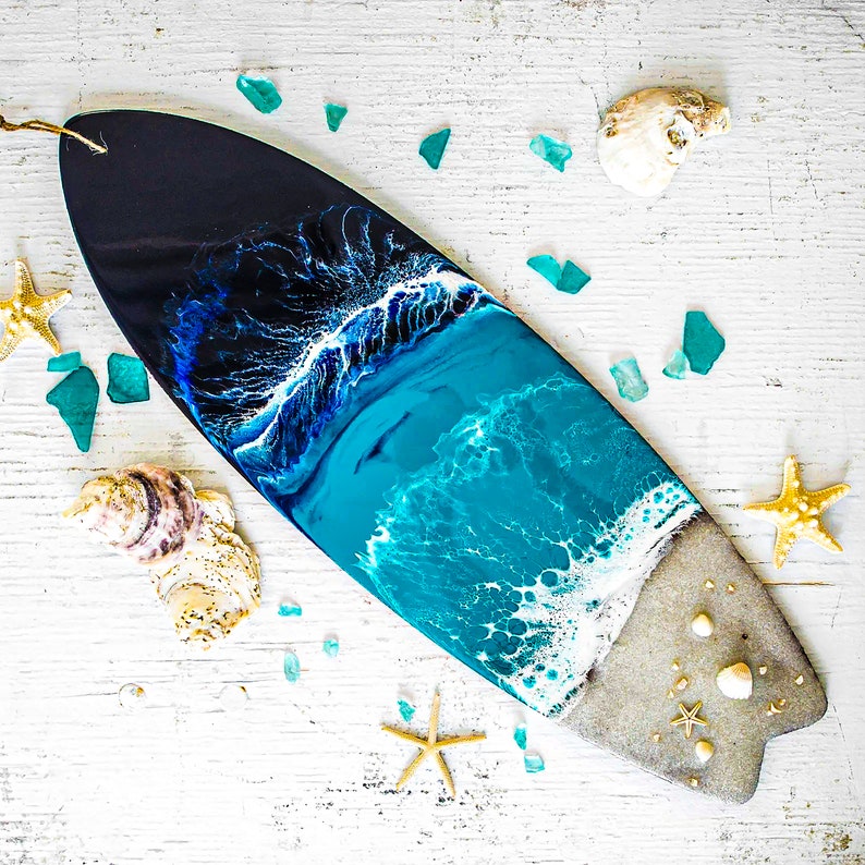 May include: A black and blue surfboard with a resin ocean wave design. The surfboard is decorated with seashells and small pieces of sea glass.