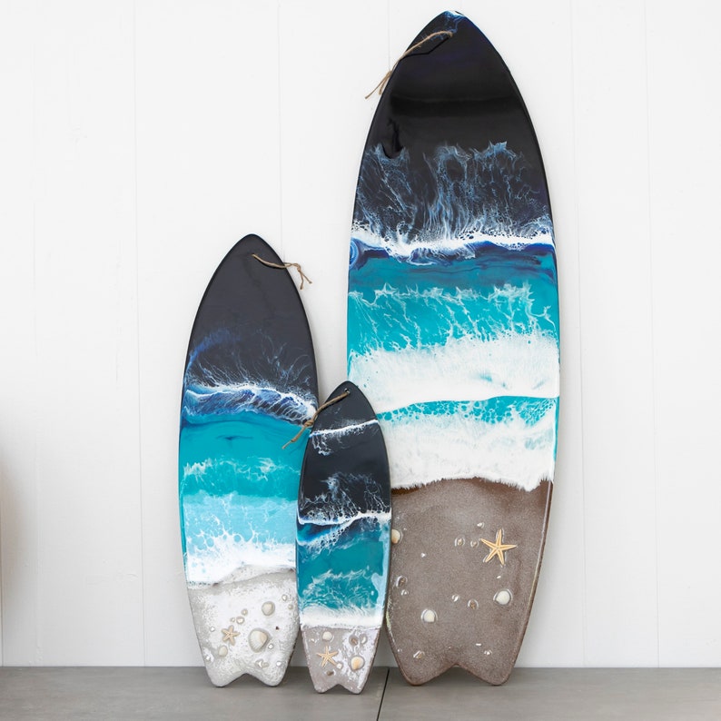 May include: Three resin surfboard wall decor pieces in different sizes. The largest surfboard is brown with white and blue waves, the medium surfboard is blue with white waves, and the smallest surfboard is blue with white waves. All three surfboards have a black top and a white sandy bottom.