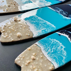 May include: Three surfboard-shaped resin art pieces with a beach theme. Each piece features a blue and white ocean wave design with a sandy beach bottom. The beach bottom is decorated with small white seashells and a starfish.
