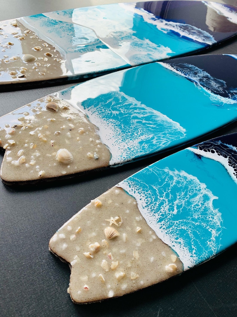 May include: Three surfboard-shaped resin art pieces with a beach theme. Each piece features a blue and white ocean wave design with a sandy beach bottom. The beach bottom is decorated with small white seashells and a starfish.