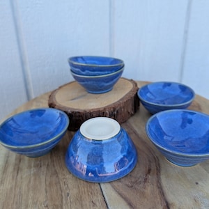 May include: A collection of small, handmade blue ceramic bowls. The bowls have a glossy finish and a slightly irregular shape, with a contrasting light-colored interior. They are arranged on a wooden surface.