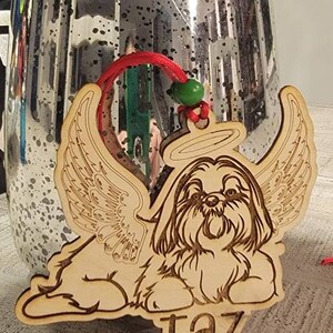 May include: Wooden ornament featuring a Shih Tzu dog with angel wings and a halo. The dog's name, "Taz," is engraved below. The ornament has a red string for hanging and a green bead.