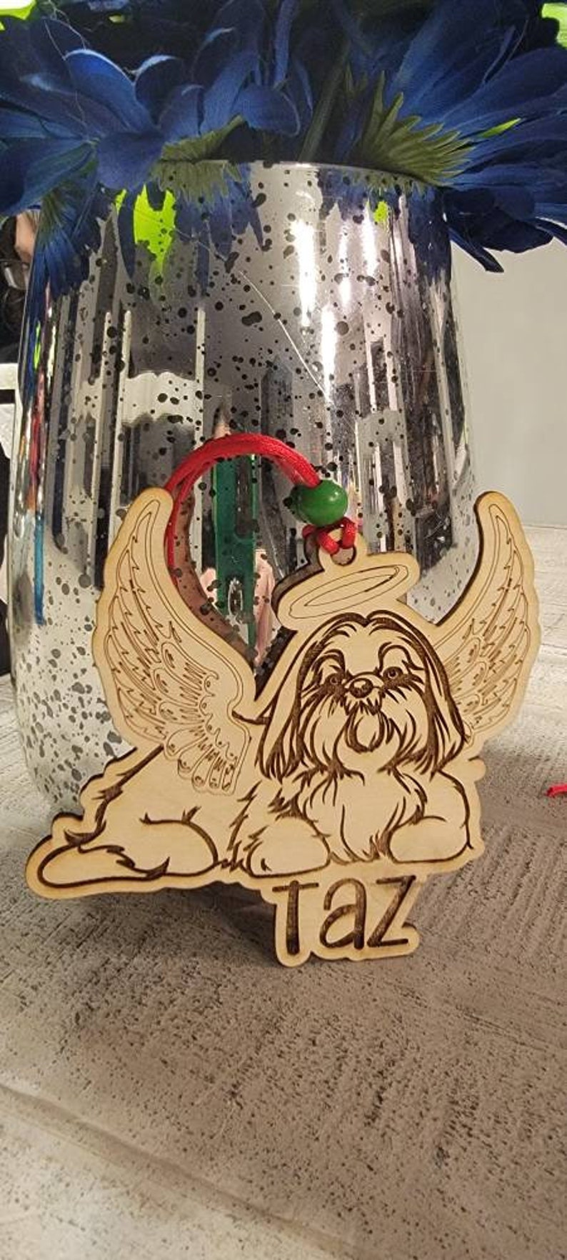 May include: Wooden ornament featuring a Shih Tzu dog with angel wings and a halo. The dog's name, "Taz," is engraved below. The ornament has a red string for hanging and a green bead.