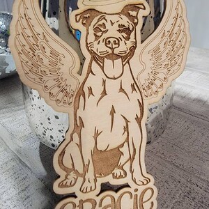 May include: Wooden ornament featuring a dog with angel wings and a halo. The dog is smiling with its tongue out. The name "Gracie" is carved at the bottom. The ornament is made of light-colored wood.