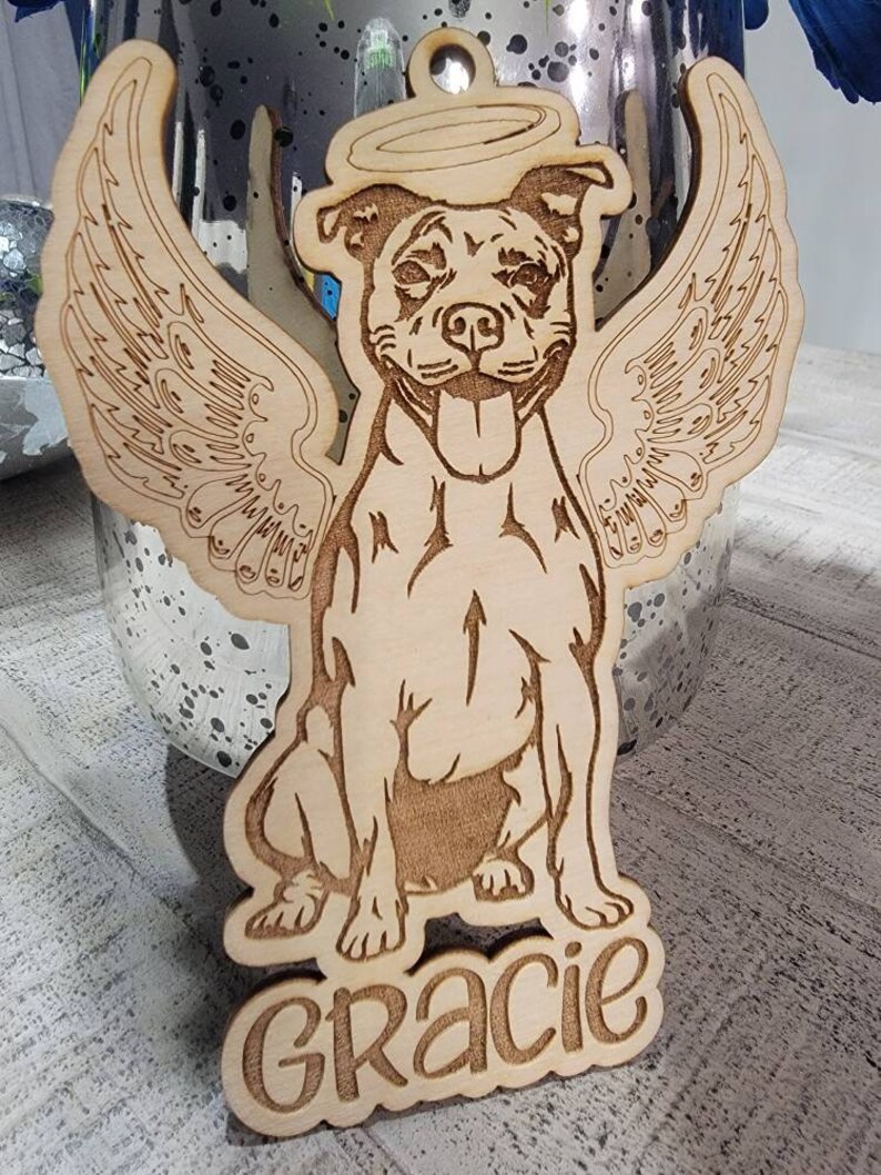 May include: Wooden ornament featuring a dog with angel wings and a halo. The dog is smiling with its tongue out. The name "Gracie" is carved at the bottom. The ornament is made of light-colored wood.