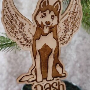 May include: Wooden Christmas ornament featuring a dog with angel wings and a halo. The dog's name, "Dash," is engraved below. The ornament hangs from a red ribbon and is set against a backdrop of evergreen branches.