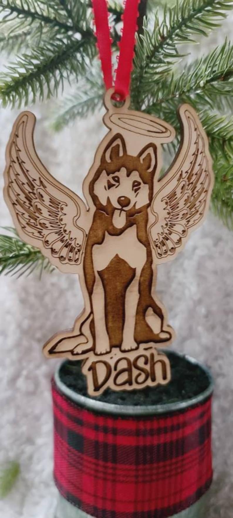 May include: Wooden Christmas ornament featuring a dog with angel wings and a halo. The dog's name, "Dash," is engraved below. The ornament hangs from a red ribbon and is set against a backdrop of evergreen branches.