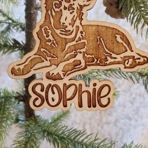May include: Wooden Christmas ornament featuring a dog design with the name "Sophie" engraved below. The ornament is suspended by a red ribbon and is displayed on a small Christmas tree. The dog is lying down.