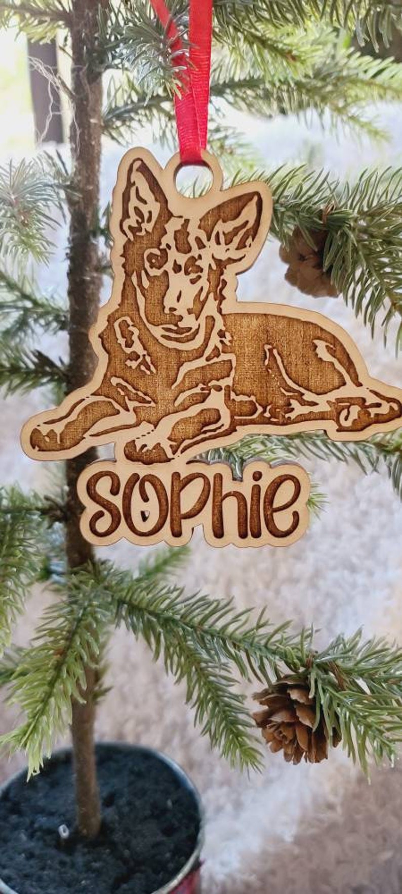 May include: Wooden Christmas ornament featuring a dog design with the name "Sophie" engraved below. The ornament is suspended by a red ribbon and is displayed on a small Christmas tree. The dog is lying down.