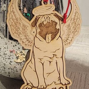 May include: Wooden pug ornament with angel wings and a halo. The pug's name, "Nugget," is engraved at the bottom. The ornament has a red string for hanging and a striped bead.