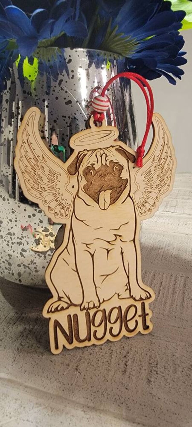 May include: Wooden pug ornament with angel wings and a halo. The pug's name, "Nugget," is engraved at the bottom. The ornament has a red string for hanging and a striped bead.