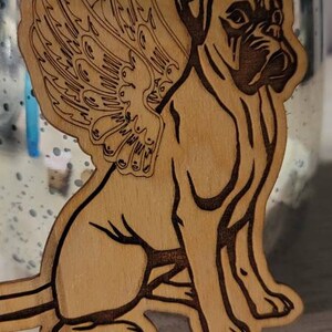 May include: Wooden ornament featuring a dog with angel wings and a halo. The dog is a boxer breed, with the name "MAC" engraved below. The ornament is made of light brown wood with dark brown etched details.