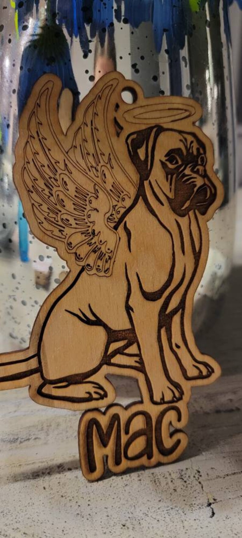 May include: Wooden ornament featuring a dog with angel wings and a halo. The dog is a boxer breed, with the name "MAC" engraved below. The ornament is made of light brown wood with dark brown etched details.