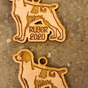 May include: Two wooden dog ornaments, each in the shape of a dog with a hole for hanging. One ornament says "Ruger 2020" and the other says "Rusty 2020". The ornaments are light brown.