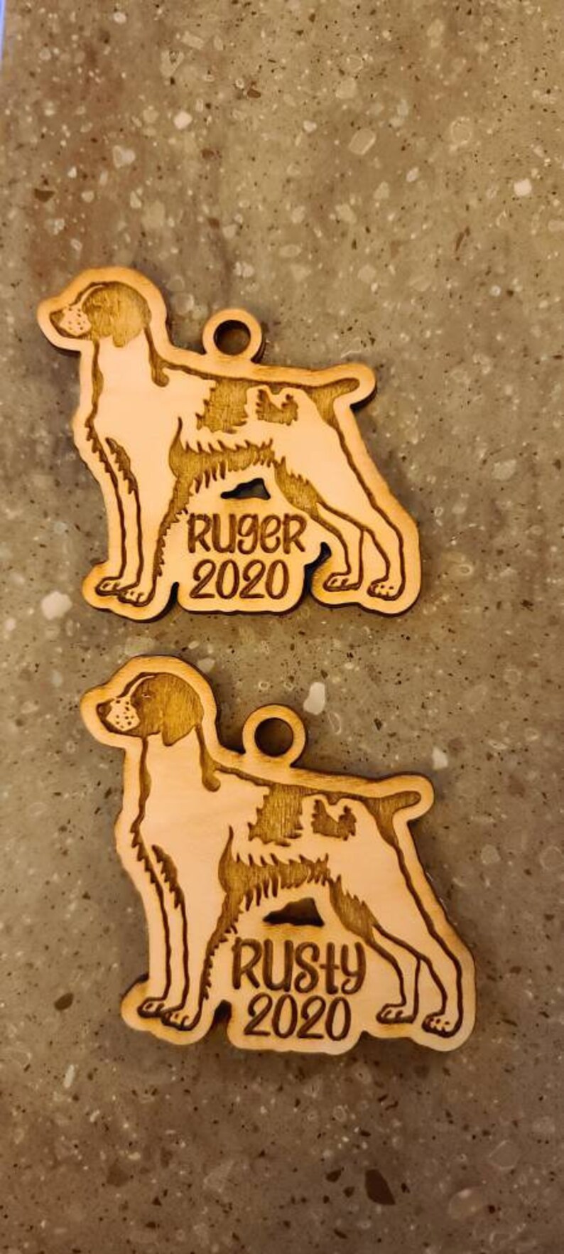 May include: Two wooden dog ornaments, each in the shape of a dog with a hole for hanging. One ornament says "Ruger 2020" and the other says "Rusty 2020". The ornaments are light brown.