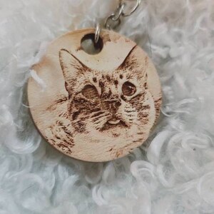 May include: A wooden keychain with a laser-engraved image of a cat's face. The cat has brown and white fur and is looking directly at the camera.