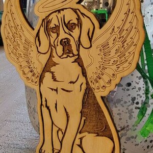 May include: Wooden ornament featuring a beagle with angel wings and a halo. The dog is engraved with detailed lines, and the name "Linus" is at the bottom. The ornament has a natural wood color and is designed for hanging.