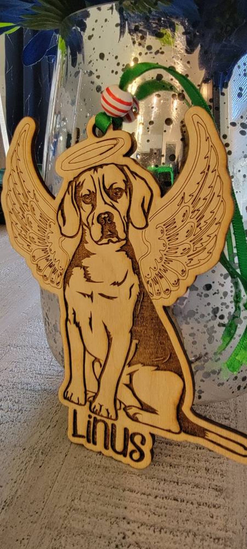 May include: Wooden ornament featuring a beagle with angel wings and a halo. The dog is engraved with detailed lines, and the name "Linus" is at the bottom. The ornament has a natural wood color and is designed for hanging.
