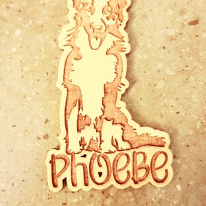 May include: Wooden dog tag featuring a detailed illustration of a Border Collie. The dog's name, "PHOEBE," is carved in a playful font below the dog. The tag has a hole for attaching to a collar.