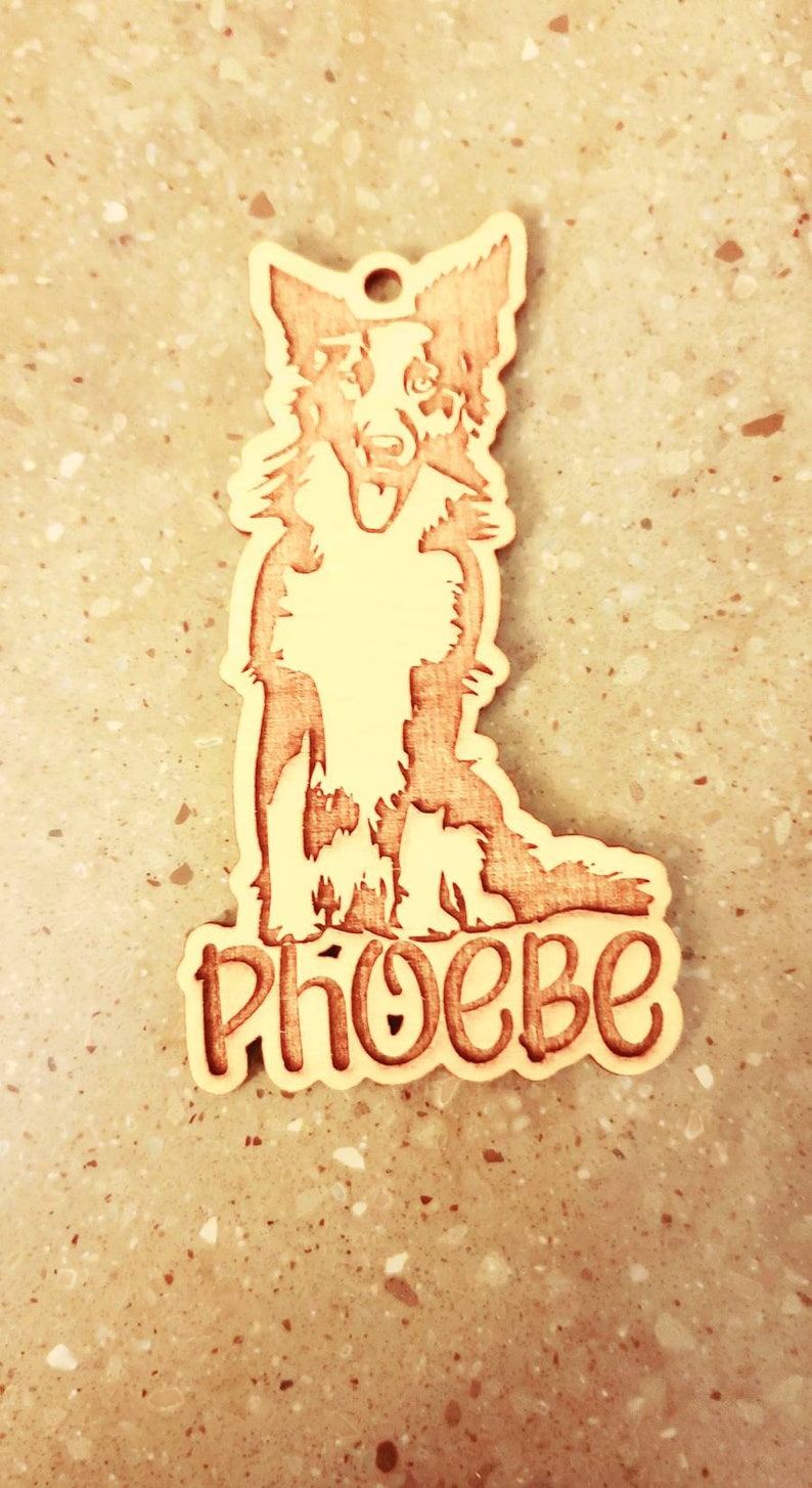 May include: Wooden dog tag featuring a detailed illustration of a Border Collie. The dog's name, "PHOEBE," is carved in a playful font below the dog. The tag has a hole for attaching to a collar.