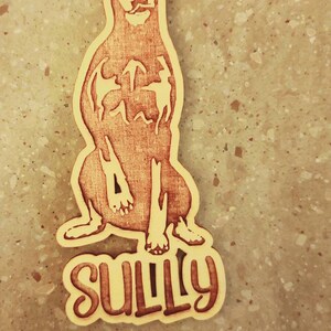 May include: Wooden dog tag featuring a Doberman Pinscher design. The dog is sitting, with the name "SULLY" in a stylized font at the bottom. The tag has a hole at the top for attachment.