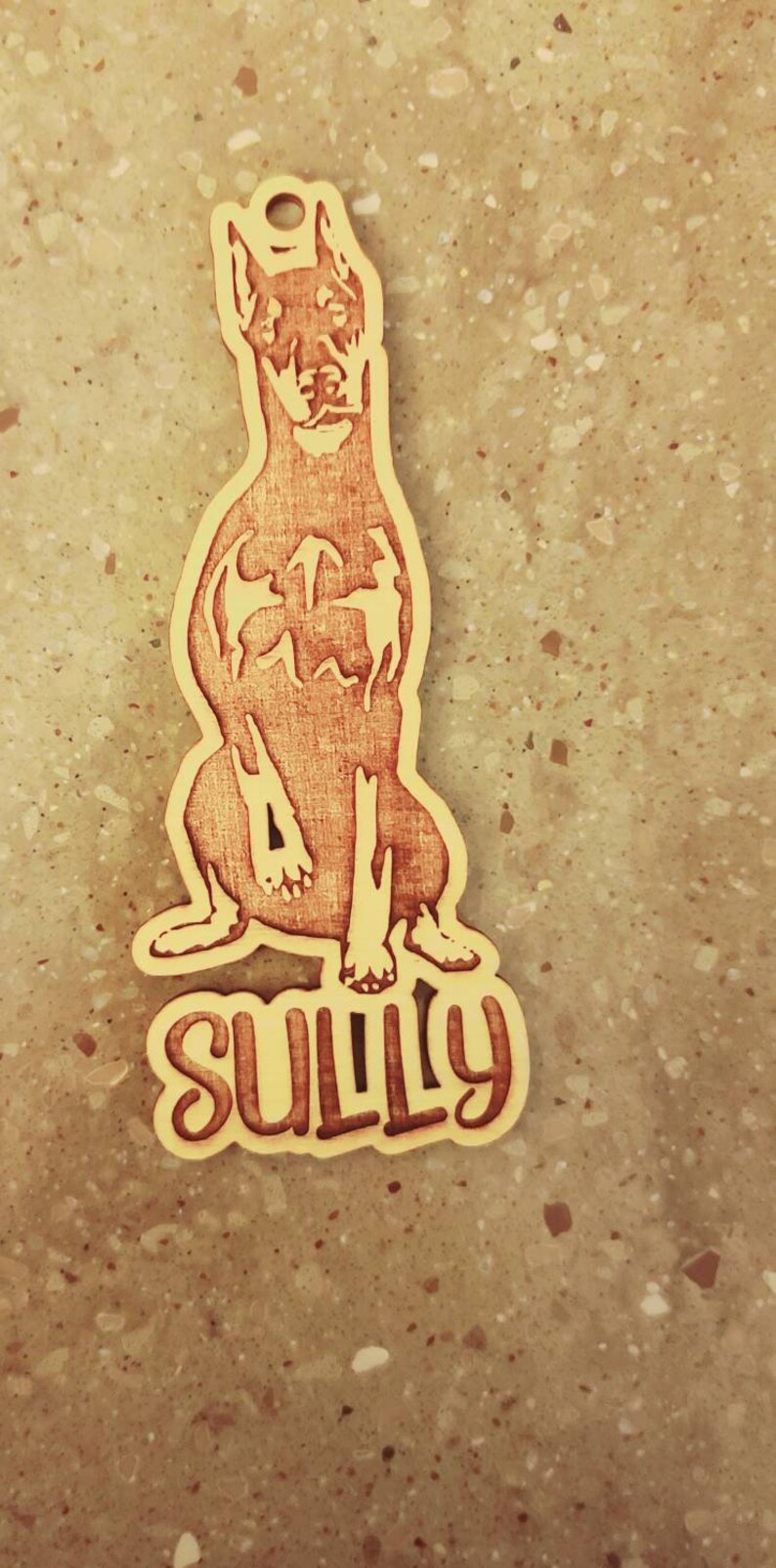 May include: Wooden dog tag featuring a Doberman Pinscher design. The dog is sitting, with the name "SULLY" in a stylized font at the bottom. The tag has a hole at the top for attachment.