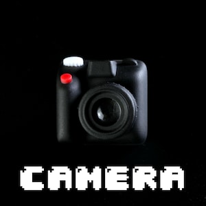 May include: A black toy camera with a red button and a white knob. The camera is on a black background with the word "CAMERA" written in white pixelated letters below it.