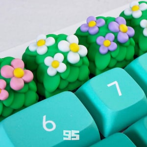 May include: A close-up of a keyboard with custom keycaps. The teal keycaps have white numbers. Decorative green keycaps shaped like bushes are adorned with pink, white, and purple flowers with yellow centres.