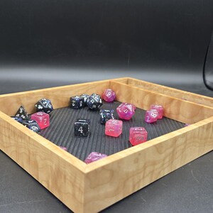 May include: A rectangular wooden dice tray with a light brown wood grain finish and a black textured base. The tray contains a collection of multi-sided polyhedral dice in various shades of pink, purple, and black with white numbering.