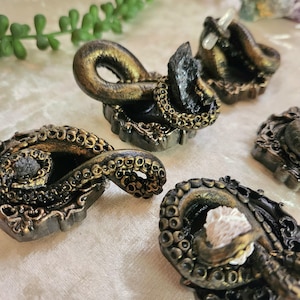May include: Several small, ornate octopus sculptures are displayed. They have a dark bronze and gold colour scheme with detailed designs. Some have a crystal or stone accent. The overall aesthetic is gothic and fantasy-inspired.