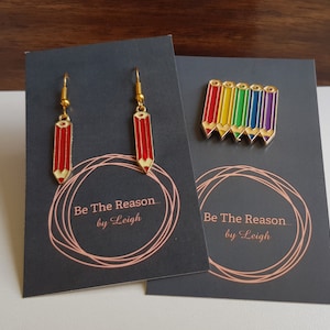 May include: A set of pencil-themed jewellery, including red pencil earrings and a rainbow pencil pin, displayed on black cards. The cards feature the text "Be The Reason... by Leigh". The earrings and pin have gold-coloured accents.