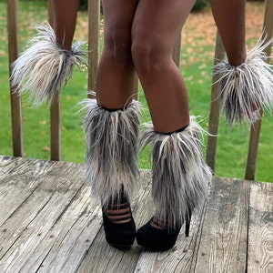 May include: A pair of black heeled boots with white and gray faux fur leg warmers. The leg warmers are made of fluffy, long-haired fur.
