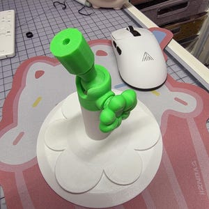 Könnte beinhalten: Ein 3D-gedruckter, beweglicher Telefonständer. Der Ständer hat eine weiße Basis mit einem Blütenblatt-Design, einen weißen zylindrischen Mittelteil und einen hellgrünen, verstellbaren Arm und Telefonhalter. Der Ständer ist für ein Telefon oder ein kleines Gerät konzipiert.