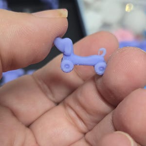 Może przedstawiać: Mała, jasnofioletowa figurka jamnika z kółkami, trzymana między palcami. Miniaturowy pies ma zaokrąglony korpus, długi grzbiet i zakrzywiony ogon. Koła są okrągłe, a uszy psa są podniesione.