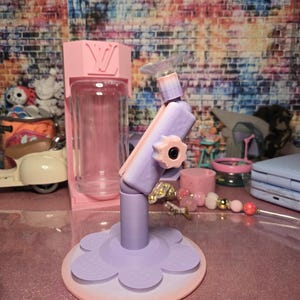 May include: A pink and purple phone stand with a flower-shaped base. The stand has a clear suction cup at the top and a pink container in the background. The background is a colorful brick wall.