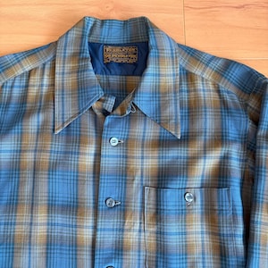 May include: A blue, tan, and brown plaid button-up shirt with a pointed collar and a chest pocket. The shirt has a label that reads "Pendleton" in gold lettering.