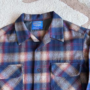 May include: A close-up of a Pendleton plaid shirt in shades of blue, red, and tan. The shirt has two chest pockets, a button-down front, and a collar. The label reads "Pendleton Made in U.S.A."