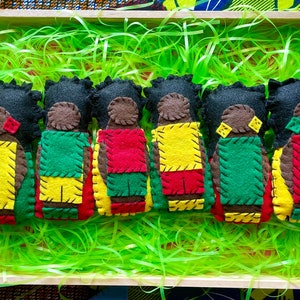 May include: Seven felt dolls with black hair and brown faces, each wearing a different color combination of red, yellow, and green clothing. The dolls are arranged in a row in a wooden box lined with green shredded paper.