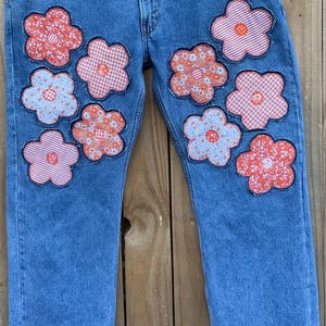 May include: Blue denim jeans with a patchwork design of various floral patterns in shades of pink, orange, and white. The flowers are sewn onto the front of the jeans.