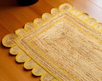 Puede incluir: Una alfombra rectangular de yute con un borde amarillo festoneado. La alfombra tiene una textura natural y tejida y un borde amarillo que enmarca el rectángulo interior. La alfombra está colocada sobre un suelo de madera cerca de una ventana.